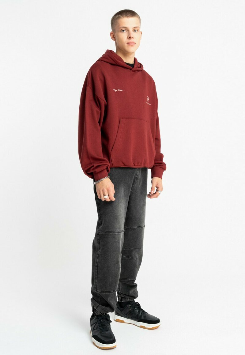 Bordowa bluza z kapturem z przednią kieszenią i logo, połączona z ciemnoszarymi dżinsami jogger z detalami przeszycia oraz czarnymi sneakersami na gumowej podeszwie.