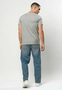 Grå bomulls-t-skjorte med korte ermer, rund hals, brukt sammen med blå denimjeans og hvite joggesko, sett bakfra.