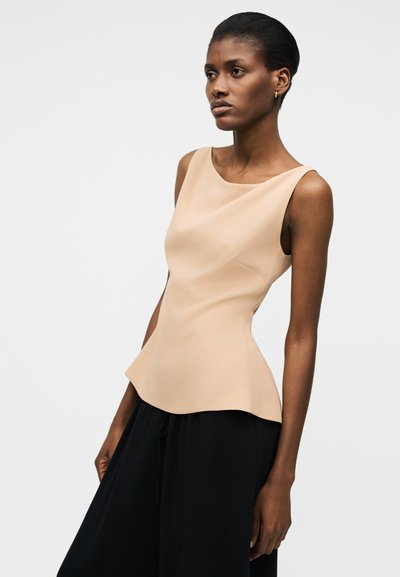 Žena tamne puti i kosa povezana unazad, nosi bež top bez rukava i široke crne hlače, na običnoj pozadini.