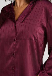 Bordowa satynowa koszula w pionowe paski, zapinana z przodu, z pojedynczą kieszenią na piersi oraz długimi rękawami z guzikami przy mankietach. Gładka faktura.