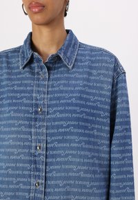 Hellblaue Jeanshemd mit einem ganzflächigen, wiederholten weißen "Tommy Jeans"-Muster, ausgestattet mit einem klassischen Kragen und silbernen Druckknöpfen.