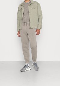 Man med en ljus khakifärgad jacka över en taupe-färgad sweatshirt och joggers, med grå och vita sneakers, stående på en enkel ljus färgad golv.