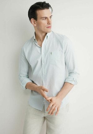 Mężczyzna w jasnooliwkowej koszuli w kratę i jasnobeżowych spodniach stoi oparty o białą ścianę, patrząc w bok z luźno złączonymi rękami.