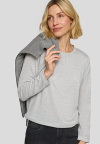 Light grey long-sleeve shirt with a round neckline and soft texture, paired with a dark grey knitted sweater draped over one shoulder.