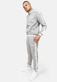 Grijze trainingsoutfit met witte strepen, ritsjack met hoge kraag, gestructureerde stof, aansluitende broek en witte sportschoenen.