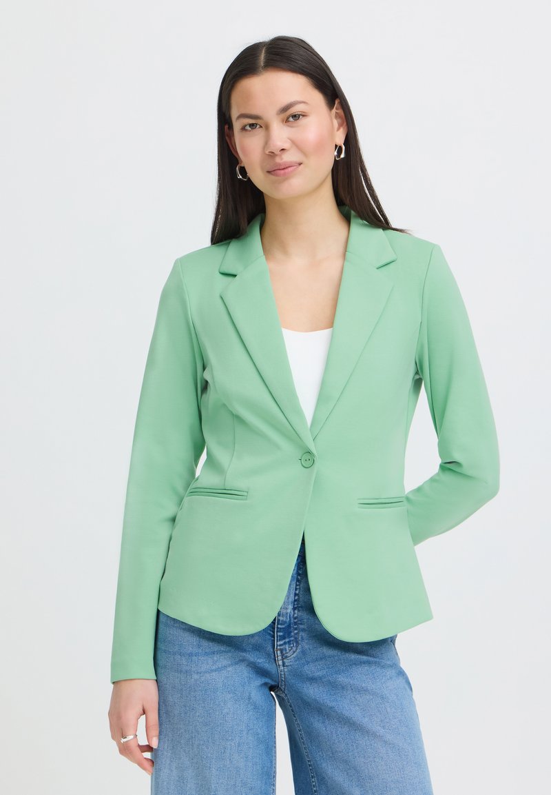 Jeune femme aux longs cheveux foncés portant un blazer ajusté vert clair, un haut blanc, un jean bleu et des boucles d'oreilles créoles, posant avec une main derrière le dos.
