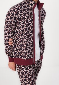 Chaqueta con cremallera y pantalones a juego en un patrón geométrico en rojo oscuro, blanco y marino, con puños y dobladillo acanalados. La textura de la tela parece suave.