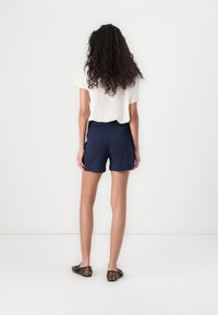 Femme aux longs cheveux bouclés portant un blouse blanche à manches courtes, un short bleu marine et des mocassins à imprimé léopard, se tenant devant un fond blanc uni.