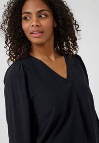 Blouse noire à manches longues avec un col en V et un détail de manches froncées. Fabriquée en tissu texturé, elle possède une poche sur le côté gauche.