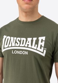 Olivinės spalvos medvilninė marškinėliai su dideliu baltu "LONSDALE" logotipu ir "LONDON" tekstu žemiau, apvaliu kaklu, trumpomis rankovėmis ir minkšta tekstūra.