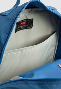 Mėlynos audinių kuprinės vidus, rodantis šviesiai pilką pamušalą ir kišenę. Yra „Levi's“ etiketė juodos spalvos su raudonais akcentais. Uždarymas su užtrauktuku.