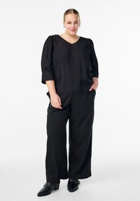 Vrouw die met één hand in haar zak staat, gekleed in een zwarte V-hals blouse, wijde zwarte broeken en zwarte puntige schoenen met gespen.