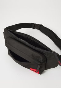 Sacoche en tissu noir avec deux compartiments zippés et une sangle ajustable, dotée de petites étiquettes rouges de la marque HUGO sur chaque tirette de fermeture.