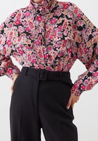 Floral patterned blouse in black, pink, and yellow, featuring a mandarin collar and long sleeves, paired with high-waisted black trousers and belt.