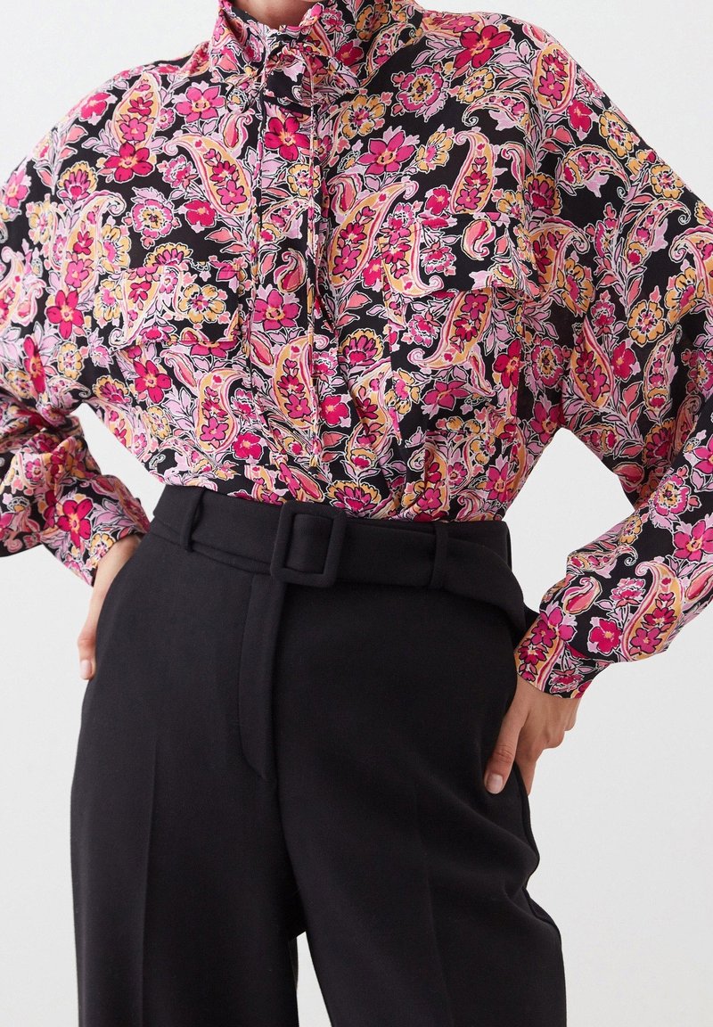 Floral patterned blouse in black, pink, and yellow, featuring a mandarin collar and long sleeves, paired with high-waisted black trousers and belt.