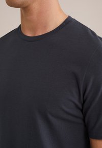 Bărbat îmbrăcat cu un tricou simplu, pe fundal închis, de culoare albastru marin, cu guler și mâneci scurte, având cusături vizibile.
