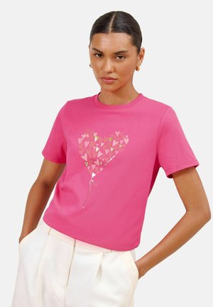 Kvinde iført pink t-shirt med ballon formet som et hjerte lavet af mindre hjerter og hvide bukser, stående med hænderne i lommerne.