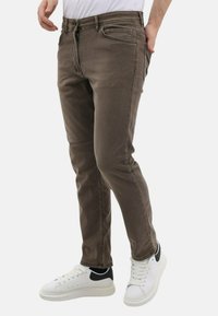 Bruine slim-fit jeans met details op de voorzakken, een ritssluiting en subtiele vervaging; gedragen met witte sneakers met zwarte accenten.