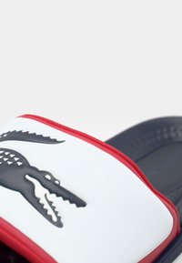 Hvid sandle med en struktureret mørkeblå sål, rød kant og et sort krokodille-logo på remmen. Glat materiale med et fladt design.