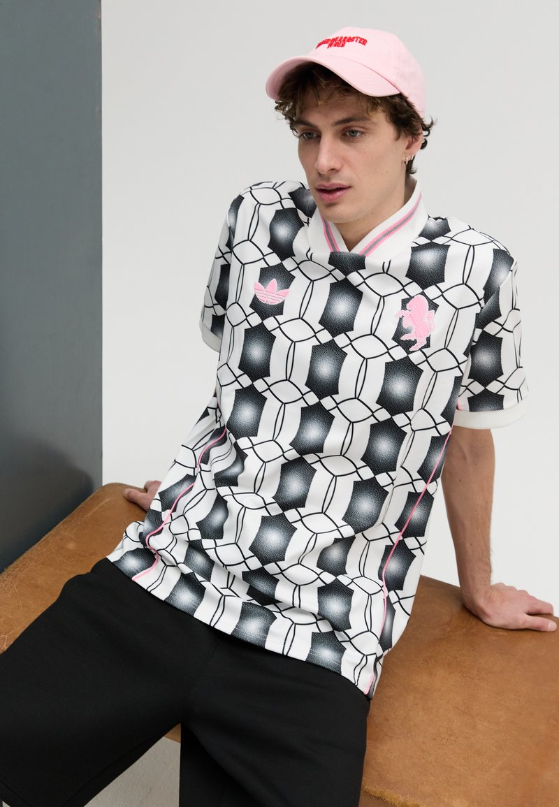 White shirt with black geometric pattern, pink accents, and Adidas logo. Black shorts. Pink cap with text. Smooth texture, relaxed fit.