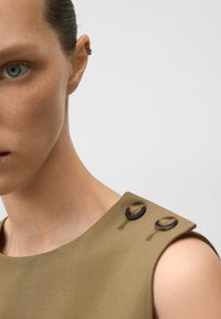 Närbild av en persons axel som bär en ärmlös beige topp med två mörka knappar på axeln och en silverörkrok.