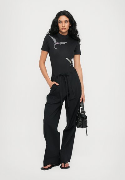Black fitted top with short sleeves featuring metallic wing designs; paired with high-waisted black wide-leg pants and a small black bag.