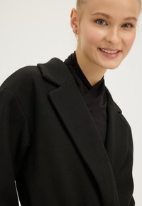 Smiling woman with light skin and blue eyes wears gold chain earrings, a black high-neck top, and a black wool coat with wide lapels.