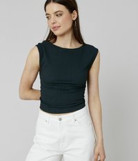 Jeune femme portant un haut texturé sans manches noir et un pantalon blanc taille haute, regardant sur le côté avec une main derrière le dos.