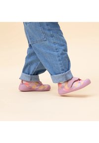 Zapatos rosas coloridos con parte superior de tela suave y un patrón geométrico, cierre de velcro y suela de goma texturizada, usados con pantalones de mezclilla enrollados.
