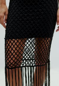 Zwarte macramé rok met een diamantnetontwerp en franjes aan de onderkant. Materiaal lijkt gestructureerd; huid is zichtbaar door de openingen.