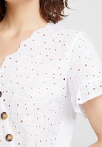 Blouse blanche avec des motifs floraux en œillets, décolleté festonné, manches courtes et boutons bruns sur le devant. Tissu doux et texturé.