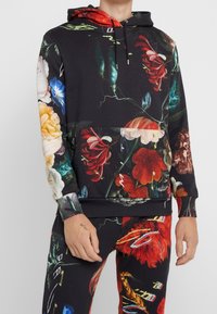 Hoodie i svart tyg med ett övergripande blommönster i livfulla färger. Inkluderar en framtill placerad ficka och ribbade ärmslut.