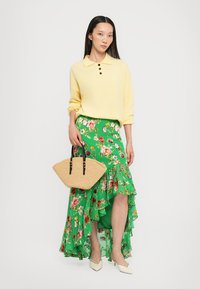 Golfino giallo lavorato a maglia con maniche corte, gonna maxi floreale verde con volant, borsa di paglia intrecciata e scarpe crema a punta.