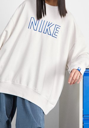 Personne portant un sweat-shirt blanc Nike oversize avec des lettres bleues, posant la main sur un meuble métallique bleu.