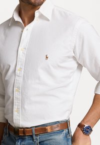 Camicetta bianca con logo di un piccolo cavallo ricamato, abbinata a jeans di denim blu e una cintura in pelle marrone. Orologio con quadrante blu e cinturino marrone.