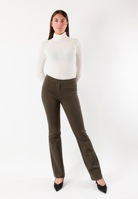 Maglia bianca a collo alto abbinata a pantaloni aderenti verde oliva scuro, con una texture liscia e una leggera forma svasata, completata da tacchi neri.