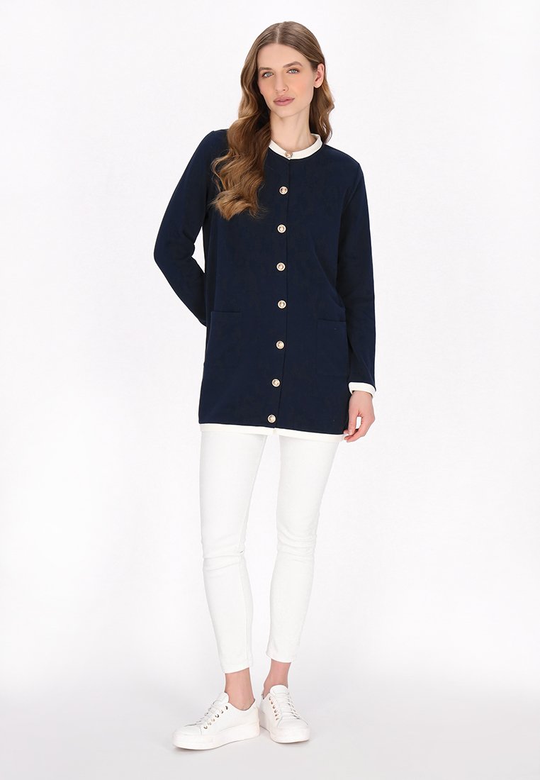 Lange marineblauwe jas met een ronde halslijn, gouden knopen en twee frontzakken, gecombineerd met witte broek en witte sneakers.