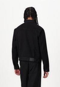 Svart denimjakke med rett passform. Har krage, knapper og en merket etikett bak på livet. Glatt tekstur og klassisk design.