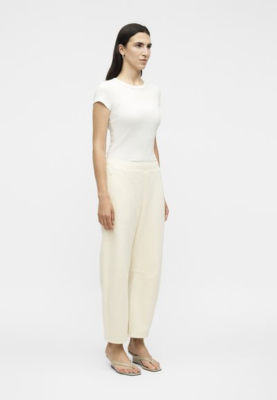 Γυναίκα που στέκεται πλαγίως, φορώντας λευκό εφαρμοστό κοντομάνικο τοπ, χαλαρά κρεμ παντελόνια και μπεζ πέδιλα με τακούνι σε μονόχρωμο φόντο.