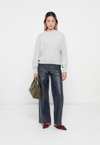 Ung kvinne står foran en hvit bakgrunn iført en grå hettegenser, vide mørke jeans, røde leiligheter, og holder en olivengrønn håndveske.
