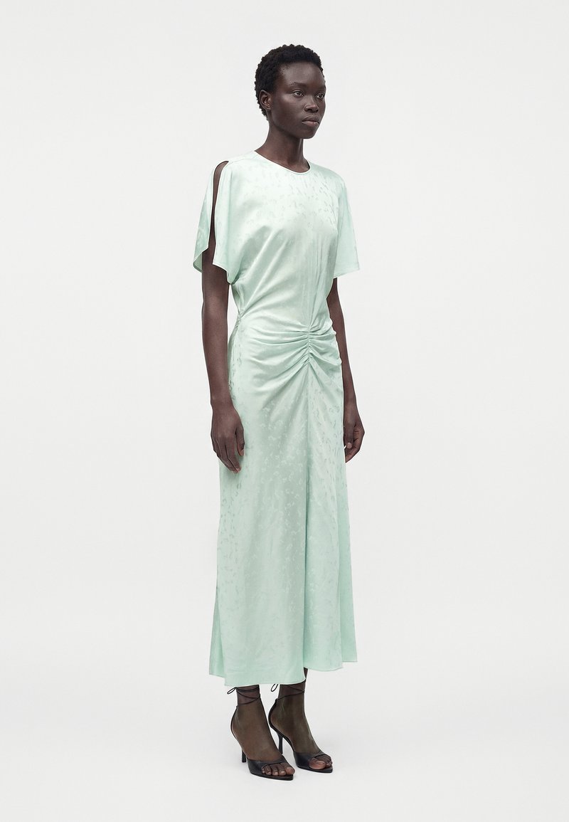 Femme portant une robe midi vert clair avec des manches fendues sur le côté et un détail froncé à la taille, associée à des talons ouverts noirs.