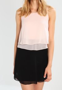 Ärmös topp i ljusrosa som är lager på lager över en svart kjol. Tyget är genomskinligt med en slät textur, med en rundad halsringning och en luftig design.