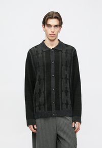 Crni i tamnosivi kardigan s uzorkom, ovratnikom, kopčanjem na gumbe i teksturiranim tkanjem. Sadrži vodoravne i okomite uzorke.