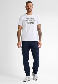 Vit bomullströja med grön grafisk tryck som står "NASAR CUP DAYTONA PETROL IND. EST. 1989". Matchas med marinblå cargobyxor och vita sneakers.