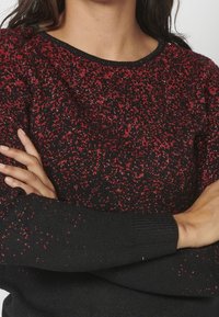 Schwarzer Pullover mit roten und metallischen Fäden, ausgestattet mit einem strukturierten Strickdesign. Lange Ärmel mit gerippten Bündchen. Runder Halsausschnitt.