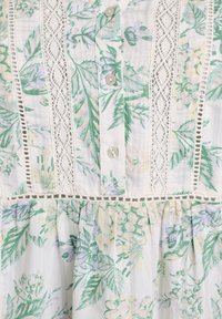 Robe florale avec des motifs botaniques verts et violets, bordure en dentelle blanche et boutons en tissu. Matériau en coton léger et texturé.