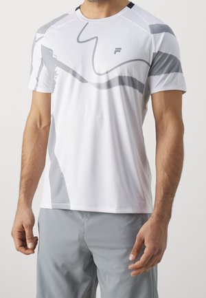 Mężczyzna ubrany w biały sportowy t-shirt z krótkim rękawem z szarymi abstrakcyjnymi wzorami oraz jasnoszare szorty, stojący na jednolitym tle.