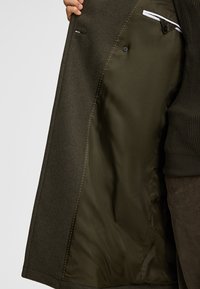 Manteau vert olive foncé avec un extérieur texturé. L'intérieur est doté d'une doublure lisse, d'un bouton et d'une poche cachée, ainsi que de détails de couture blanche.
