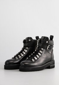 Bottes noires en cuir avec œillets argentés, fermeture éclair latérale, sangle à boucle et semelle épaisse crantée, posées sur une surface claire.