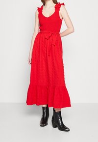 Punainen midi-mekko, jossa on röyhelöolkaimet, teksturoitu vartalo ja lanteilta levenevä kerrostettu helma. Yhdistetty mustiin nilkkasaappaisiin, joissa on krokotiilikuosia.